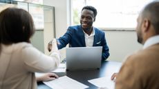 A man in a suit smiles and shakes hands with the woman across the table from him.