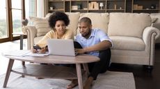 A couple sit on the floor in front of their sofa and look at a laptop together.