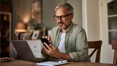 An older man looks at his phone while sitting at his dining room table in front of his open laptop.